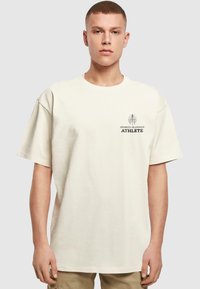 Krēmkrāsas pārāk liels T-krekls, kas izgatavots no kokvilnas. Tam ir īsas piedurknes un grafiskais prints ar uzrakstu "SPORTA SEZONA SPORTISTS" tumšā krāsā.