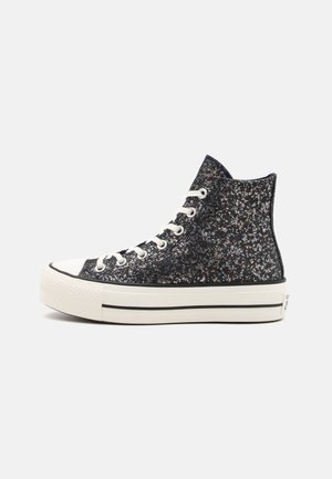 Zapatillas altas con tela negra brillante, suela de goma blanca y detalles en negro. Presentan cordones en la parte delantera y punta redondeada.