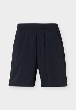 Personne portant un short de sport noir, des bottes de randonnée noires avec des lacets blancs, des chaussettes blanches et un haut noir à manches longues, debout sur un fond blanc.