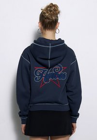 Granatowy hoodie wykonany z miękkiego materiału, z czerwonymi i niebieskimi haftowanymi literami "Bohaterowie" oraz wzorem w gwiazdę na plecach i kontrastowymi przeszyciami.
