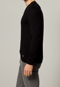 Maglione nero a maniche lunghe in maglia con scollo a V, design aderente e polsini a costine. Etichetta visibile all'orlo. Indossato con pantaloni grigi in vista laterale.