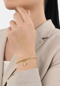 Bracciale in oro con charm a forma di farfalla dalle ali dettagliate. La mano è posizionata vicino a un blazer di colore neutro.