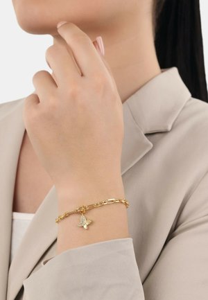 Golddrahtarmband mit einem Schmetterlingsanhänger mit detaillierten Flügeln. Die Hand ist in der Nähe eines blass gefärbten Blazers positioniert.