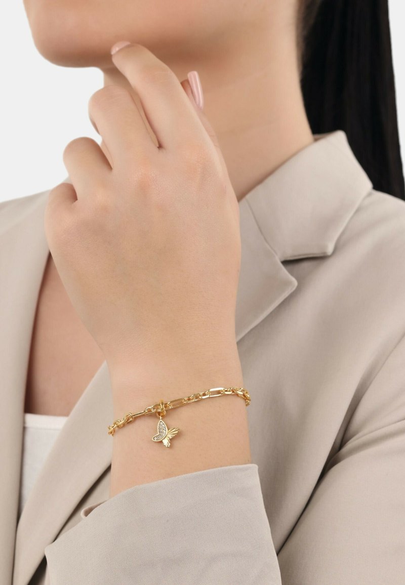 Bracciale in oro con charm a forma di farfalla dalle ali dettagliate. La mano è posizionata vicino a un blazer di colore neutro.