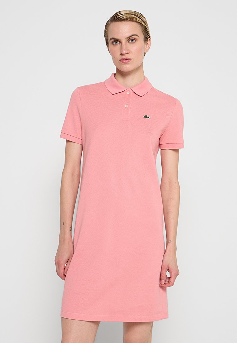 Robe polo en coton rose clair avec un col et des manches courtes. Présente un petit logo vert sur la poitrine gauche.