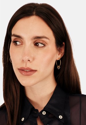 Gold hoop earrings with a heart-shaped accent. Model wears a sheer black collared shirt. Smooth skin with natural makeup.