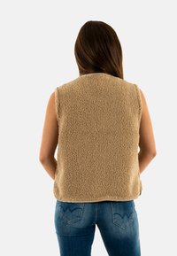 Gilet in pile color cammello con superficie texture e morbida. Design senza maniche e orlo arrotondato. Abbinato a jeans in denim con un design distintivo della tasca posteriore.