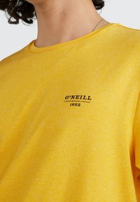 Camiseta amarilla de manga corta hecha de una tela suave. Presenta un pequeño logo negro de "O'NEILL" y el año "1952" impreso en la parte delantera.