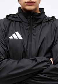 Άτομο που φοράει αθλητικό μαύρο μπουφάν με φερμουάρ και ψηλό γιακά, με λευκό λογότυπο τριών ρίγων στο αριστερό στήθος, με σταυρωμένα χέρια.