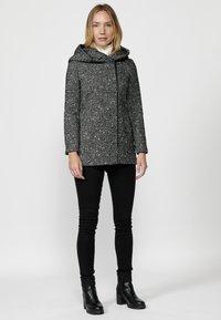 Šedý kabát s textúrovaným, bodkovaným vzorom a prehnanou kapucňou. Obsahuje predný zips a voľný strih. Nosený s čiernymi skinny jeans a čižmami.