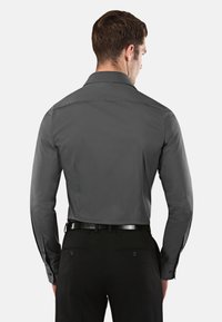 Camicia grigia a maniche lunghe con colletto classico e polsini con bottoni, caratterizzata da una texture liscia. Indossata con pantaloni neri su misura e una cintura nera.
