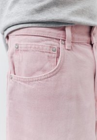 Gros plan sur la taille d'un jean en denim rose clair avec une poche avant, passants de ceinture, et une partie d'une chemise grise rentrée.