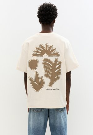 Beige t-shirt med store brune planteformer på ryggen, med en syet kant og teksten "living pattern" nederst.
