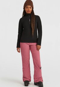 Schwarzes, langärmliges Oberteil mit Reißverschluss, kombiniert mit pinkfarbenen Schneehosen mit strukturierter Oberfläche. Braune Mütze und schwarze Schneestiefel vervollständigen das Outfit.