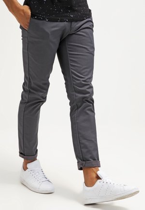 Uomo che indossa pantaloni slim fit grigio scuro con risvolti arrotolati e sneakers bianche, in piedi con una mano in tasca su sfondo bianco.