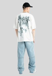 Biała, oversize'owa koszulka z szarym abstrakcyjnym wzorem na plecach, w zestawieniu z luźnymi niebieskimi dżinsami i czapką w kolorze czarnym.