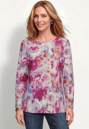 Femme aux cheveux blonds mi-longs portant un haut à manches longues multicolore à motifs floraux et un jean bleu foncé, souriant légèrement.