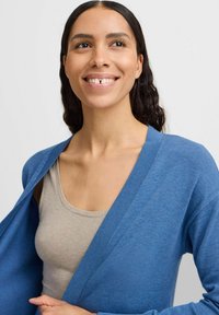Blauwe cardigan van zacht materiaal met een relaxte pasvorm, voorzien van een diepe v-hals en ribbels aan de randen, gedragen over een grijze tanktop.