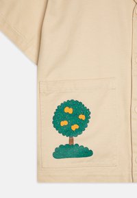 Camicia beige a maniche corte con un motivo di un albero verde con frutti gialli e una base texture sulla tasca sinistra. Tessuto liscio.
