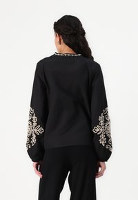 Blusa in tessuto nero con maniche lunghe, caratterizzata da un intricato ricamo floreale in crema sui polsini e una cucitura decorativa sul collo.