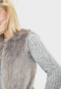 Blond person som bär en mjuk grå faux pälsväst över en strukturerad grå stickad tröja mot en ljus, neutral bakgrund.