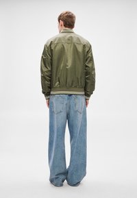 Oliveiroheleroheline pommitaja jope soonikkoelise krae ja mansettidega, kombineeritud lahtiste, helesiniste teksapükstega, nähtavad tagataskud.