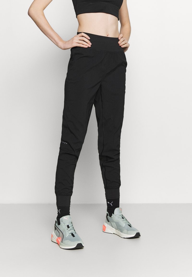 Zwarte sportieve joggers met een taps toelopend ontwerp, elastische tailleband en subtiele merkdetails. Gecombineerd met grijze sneakers en zwarte sokken.