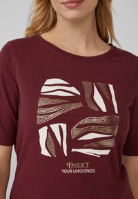 T-shirt bordeaux avec un design géométrique imprimé en blanc et or portant le texte "Accepte ton unicité." Matière en coton, manches courtes.