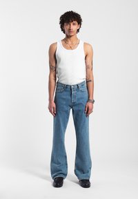 Valkoinen ribattu hihaton paita yhdistettynä vaaleansinisiin leveälahkeisiin farkkuihin, joissa on klassinen viisi taskua -malli ja hienovarainen haalistuminen. Mustat kengät ja asusteet näkyvät.