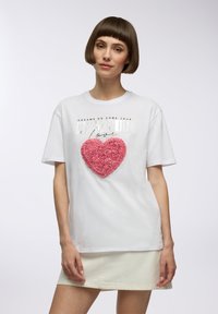 T-shirt bianco a maniche corte con un'accenno a forma di cuore rosa e testura, e testo metallizzato che legge "I SOGNI DIVENTANO REALTÀ L'AMOUR love."