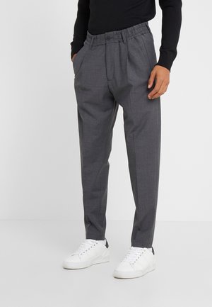 Pantaloni grigi plissettati con cintura elastica, indossati con sneakers bianche e una maglia nera a maniche lunghe, mani casualmente nelle tasche.