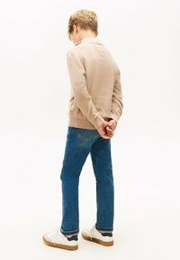 Jongen in beige trui, blauwe jeans en witte sneakers staat met zijn handen achter zijn rug gekleed, met zijn rug naar de camera toe op een witte achtergrond.