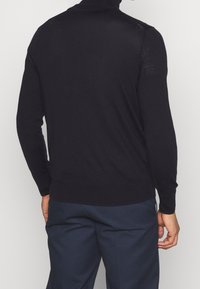 Schwarzer Strickpullover mit langen Ärmeln und hohem Kragen. Glatte Textur, taillierter Schnitt. Getragen mit dunkelblauen Hosen, Rückansicht zu sehen.