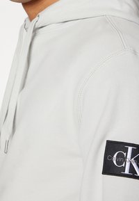 Ljusgrå hoodie med en texturerad yta, med en svart broderad Calvin Klein-logotyp på vänster arm. Dragsko i huvan.