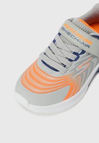 Chaussure de sport grise avec des accents orange et bleu, tige en maille, lignes texturées sur le côté et semelle blanche. Présente une sangle de marque sur le dessus.
