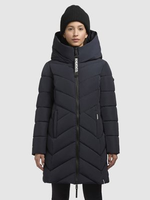 Navyblauwe pufferjas met een hoge kraag, chevronpatroon en een ritssluiting aan de voorkant. Vergezeld door een zwarte gebreide beanie.