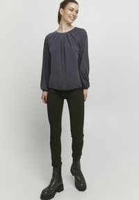 Šedá halenka s dlouhými rukávy a nabíraným detailem v oblasti krku, v kombinaci s tmavě zelenými slim kalhotami a tmavými kotníkovými botami. Hladká textura látky.