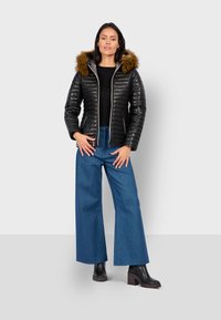 Prošívaná černá péřová bunda s kapucí podšitou kožešinou, doplněná širokými džíny. Model stojí s rukama na bocích a má na sobě černé kotníkové boty.