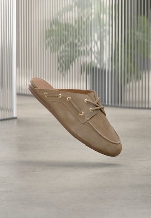 Mocasín slip-on de ante marrón claro con detalles de cordones, presentado flotando sobre un suelo gris claro y un fondo interior desenfocado.