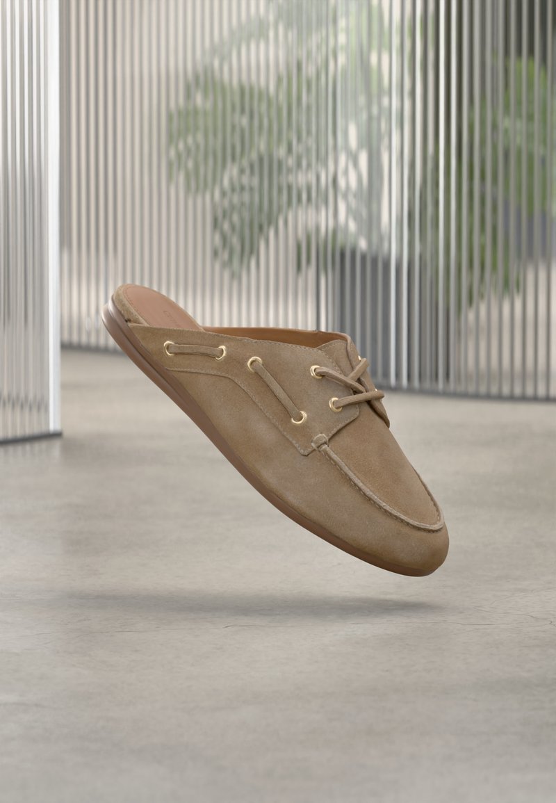 Ελαφρώς καφέ μοκασίνι από σουέτ με λεπτομέρειες κορδονιών, παρουσιασμένο να αιωρείται πάνω από ένα ανοιχτό γκρι δάπεδο και ένα θολό εσωτερικό φόντο.