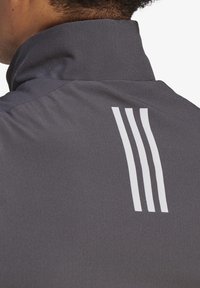 Chaqueta deportiva gris con un alto collar y tres rayas verticales blancas en la parte posterior, con un acabado de tela suave y texturizada.