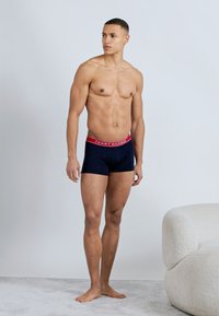 Άνδρας σε καλή φυσική κατάσταση, όρθιος, ξυπόλητος, φορώντας σκούρου μπλε ανδρικά εσώρουχα boxer με κόκκινη ζώνη μέσης και λευκό λογότυπο "Tommy Hilfiger", δίπλα σε μια καρέκλα με κρεμ ταπετσαρία.