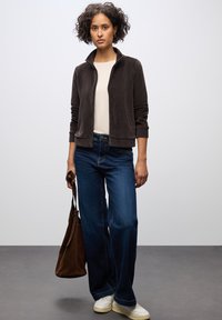 Mörkbrun kordjacka med dragkedja över en ljus t-shirt, kombinerad med vida blå jeans och vita sneakers. Brun axelväska i handen.