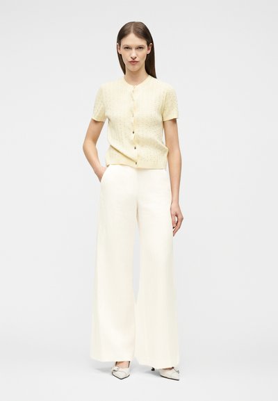 Γυναίκα όρθια με τα χέρια στις τσέπες, φοράει ανοιχτό κίτρινο κοντομάνικο πουκάμισο με κουμπιά, φαρδιά λευκά παντελόνια και λευκά μυτερά παπούτσια.