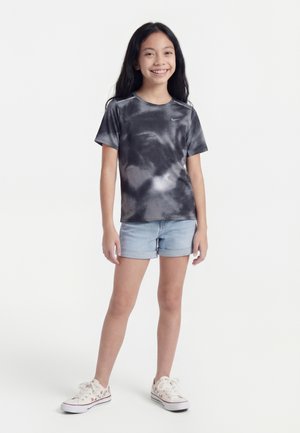 Fille souriante debout, portant un t-shirt Nike tie-dye noir et gris, un short en jean clair et des baskets blanches à motifs floraux sur un fond blanc.