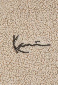 Mäkká béžová flísová tkanina s lesklým čiernym kovovým kurzívnym logom značky "Kanna" umiestneným v strede povrchu.