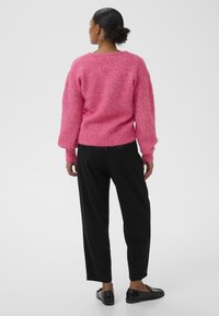 Suéter rosa de punto con mangas voluminosas, dobladillo y puños acanalados, combinado con pantalones negros de corte relajado y zapatos negros. Diseño sencillo, textura suave.
