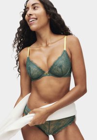 Soutien-gorge en dentelle verte avec des bretelles jaunes, présentant un design en forme de V et des bords festonnés. Culottes assorties en dentelle verte avec des détails similaires.