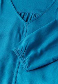 Tessuto turchese con un motivo texture, design a V e dettaglio arricciato in elastico sul polsino delle maniche e sullo scollo.