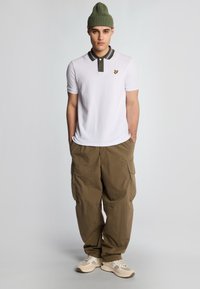 Hvid polo shirt med en mørkegrøn krave og logo, parret med løst siddende olivengrønne cargo-bukser og beige sneakers. Personen bærer en grøn beanie.